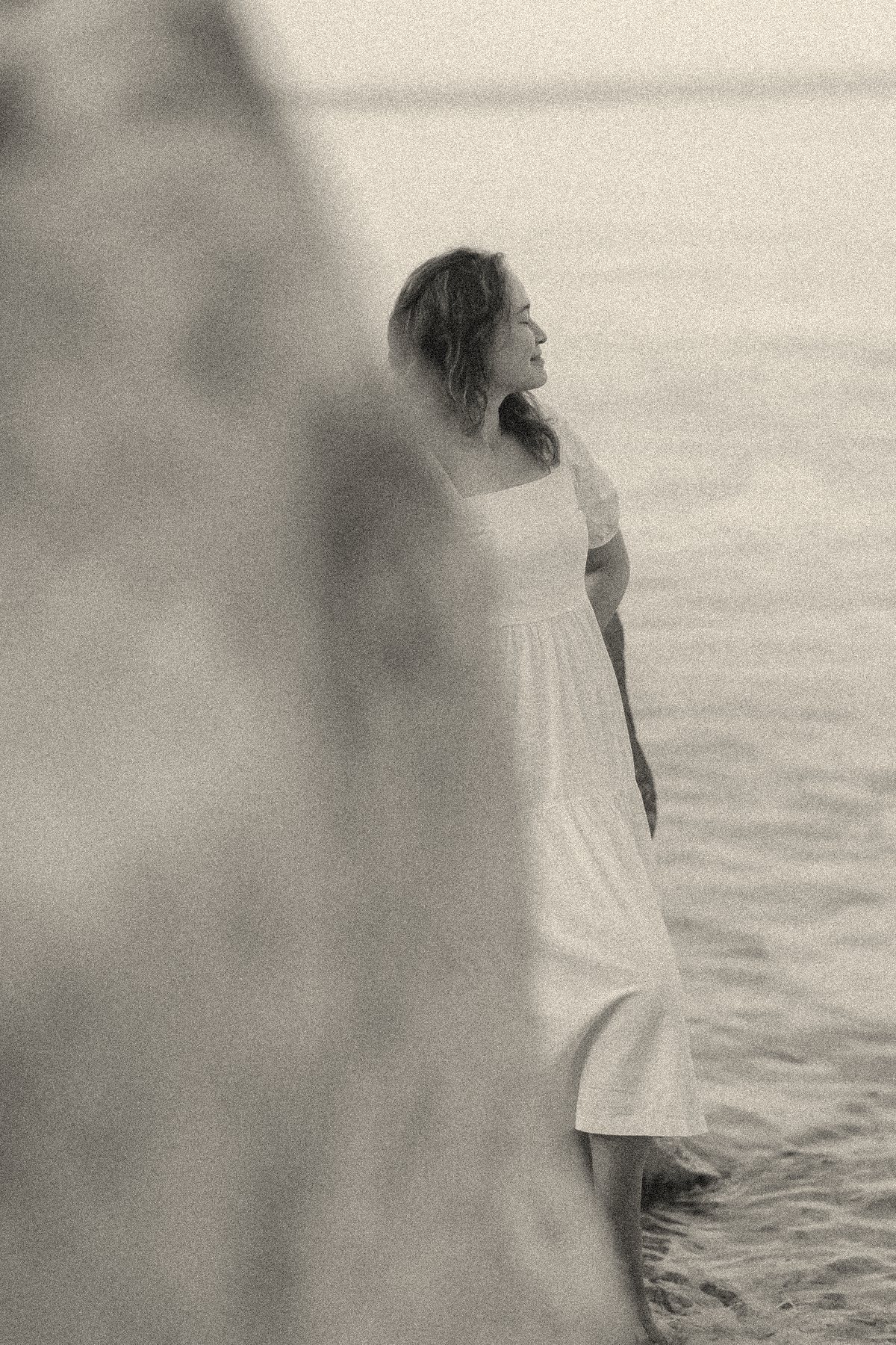 Aliya in a white dress by the sea, seen past a blurred rock